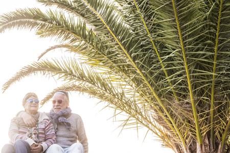 Happy old senior caucasian couple hug and stay in love together in retired lifestyle under a tropical palm - white clear sky in background - fashion people conceptの写真素材