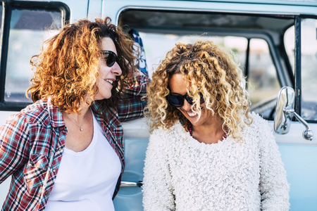 Happy people outdoor - couple of curly cute middle age adult women friends enjoy together the trip with old vintage blue van in background - laudh and happiness for hipster fashion femalesの写真素材