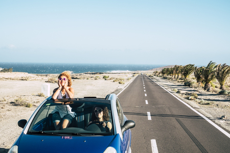 Long way road and friends travel together in freedom and independence concept with couple of young women driving for vacation - happiness and joyful - blue ocean and sky in backgroundの写真素材