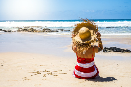 Summer holiday vacation at the beach in paradise beautiful resort people concept with nice woman with tourist hat viewed from back enjoying the sand and with sun designed on it- sea viewの写真素材