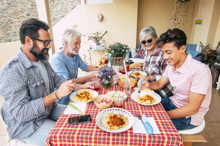 Family people outdoor lunch together on a red table - mixed ages generations enjoy and have fun with italian pasta on the terrace at home - father grandfather and sonの写真素材
