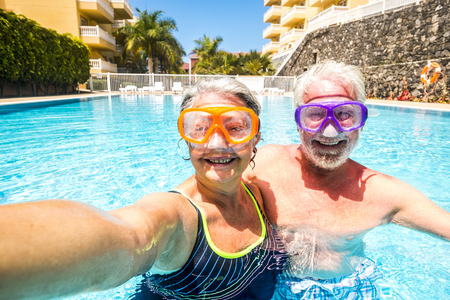 Happy cheerful people old senior man and woman have fun together in the summer swimming pool activity taking selfie pictures with scuba masks on the face for funny outdoor leisure activity in hotel holiday vacationの写真素材