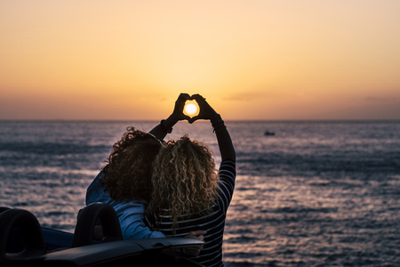 Romantic friendship people concept with two curly lady viewed from back doing hearth love sign with hands to celebrate the summer holiday vacation travel in front of the beauty of nature oceanの写真素材
