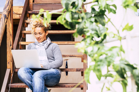 Beautiful middle age young caucasian woman working at the laptop computer internet connected outdor home sit down on a wooden stair - nice lifestyle for business activityの写真素材