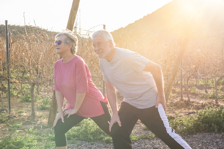 Happy active couple of old senior cacuasian people man and woman do fitness stretching for healthy lifestyle outdoor leisure activity - sunlight in backlight during sunset in country sideの写真素材