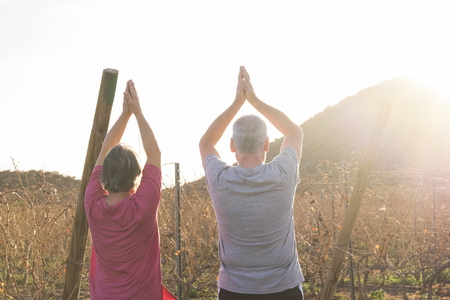 Happy active couple of old senior cacuasian people man and woman do yoga position for healthy lifestyle outdoor leisure activity - sunlight in backlight during sunset in country sideの写真素材