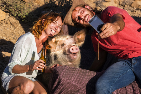 Happy alternative nature animal lover couple of cheerful people enjoy and have fun taking selfie picture with a funny pig in friendship - different lifestyle for millennials outdoorの写真素材