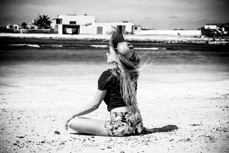 Black and white romantic and motivational portrait of beautiful blonde girl viewed from back sitting on the sand at the beach with sea and white houses in background - freedom and happiness peopleの写真素材