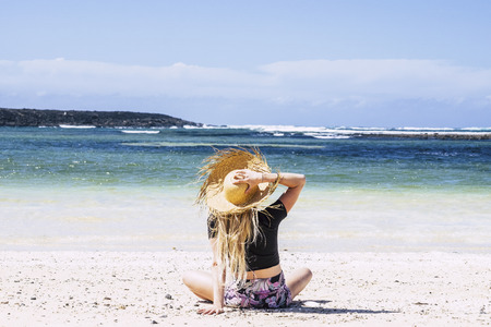 People enjoying summer holiday vacation concept with beautiful blonde babe sitting on the sand at the beach looking and feeling the blue sky and ocean in front - outdoor nature and tropical placeの写真素材