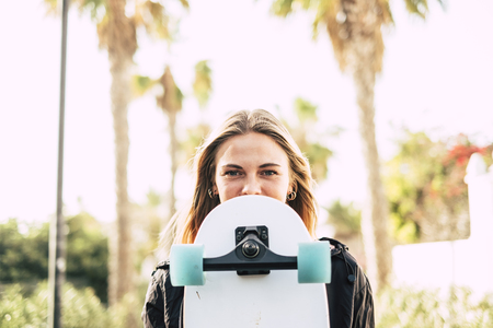 Modern cheerful beautiful blonde girl portrait with skateboard covering part of the pretty face - young millennial people outdoor concept enjoying lifestyle and sunny daysの写真素材