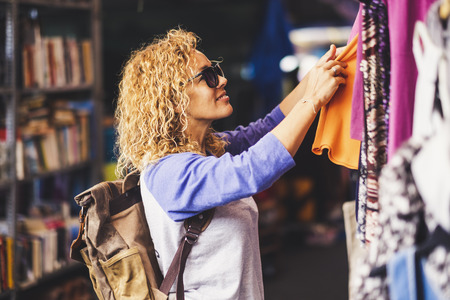 Cheerful traveler young blonde curly woman looking and choosing clothes at the used market during alternative vacation - shopping lifestyle concept for nice peopleの写真素材