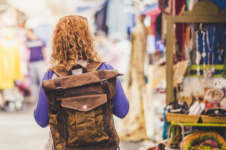 Blonde curly hair woman backpacker traveler viewed from rear at used market enjoying the shopping and alternative vacation - leather backpack view for travel conceptの写真素材