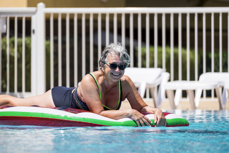 Happy cheerful caucasian people old senior woman lay down on coloured trendy lilo enjoying the swimmin pool outdoor leisure activity in summer time for vacation or relax in the cityの写真素材