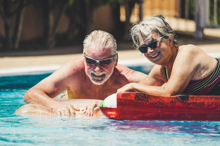 Happy joy cheerful people adult senior couple have fun in the swimming pool with trendy coloured lilos mattress on a blue water in hotel resort for summer holiday vacation lifestyleの写真素材