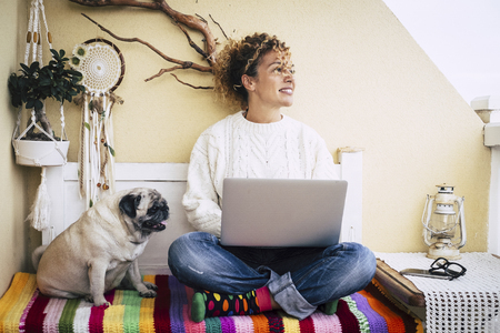 Modern couple with pretty curly woman and funny dog pug sitting on the beanch at home working with a laptop - coloured blanket and girl with technology leisure activity - millennial and love for animalsの写真素材