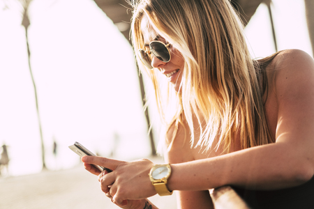 Cheerful blonde chatting at mobile phone. Smiling beautiful girl on sunset. - happy people celebrate with friendsの写真素材