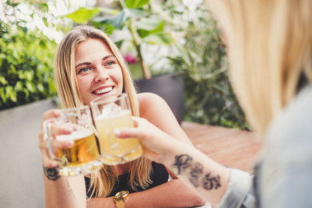 Cheerful blonde with friend at the bar drinking beer and celebrating in friendship toasting the glasses and having fun together - happy people celebrate with friendsの写真素材