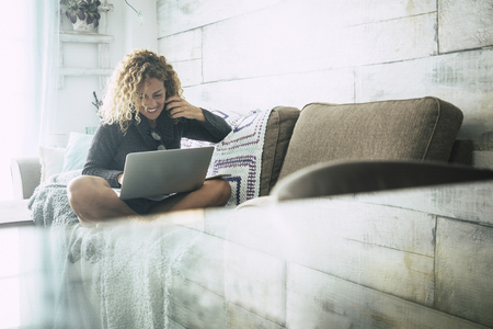 Curly woman with gray sweater sitting on brown sofa and blue blankets relaxes looking at the computer on an autumn day. Technology concept for independent people enjoying at homeの写真素材