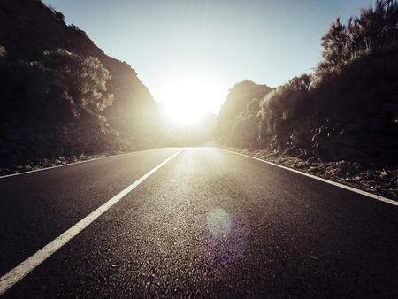 Long way road at the mountain with sun in front and sunlight effect - ground point of view with black asphalt and white lines - driving and travel conceptの写真素材