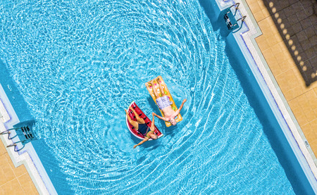 Aerial top vertical view of people old senior couple enjoying the retired lifestyle lay down with trendy coloured lilos inflatable mattress on a blue water clear swimming pool in resort hotel summer vacationの写真素材