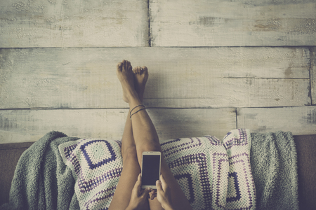 Woman lying on the couch with legs resting on the wall relaxed looking at the phone - concept of girl alone at home with technology - grey vintage color styleの写真素材