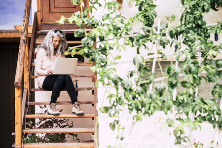 Aged beautiful woman with grey white lopng hair working at the computer laptop outside home sitting on a wooden stairs - alternative diversity people with technology - silver society conceptの写真素材