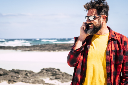 Cheerful happy hipster man with beard and sunglasses speaking at the phone with friends in outdoor beuaitufl place leisure activity - summer vacation concept with beach and ocean and sky in backgroundの写真素材