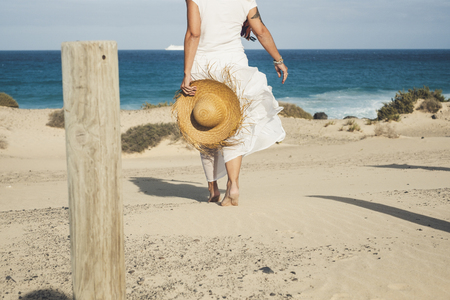 Elegant fashion lady in white dress and natural hat walking to the beach barefoot enjoying the summer holiday vacation day in tropical place - blue sea and sky horizon in background - free happy peopleの写真素材