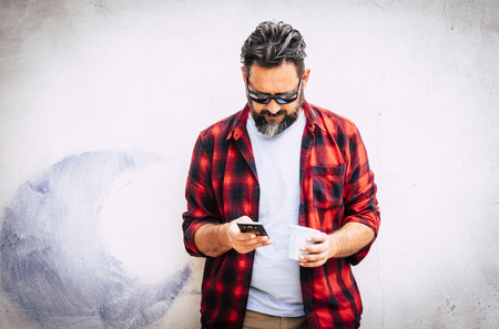 Hipster young caucasian man with beard and red shirt looking at the phone for messages or friends call - technology for senior people outdoor - white wall in backgroundの写真素材