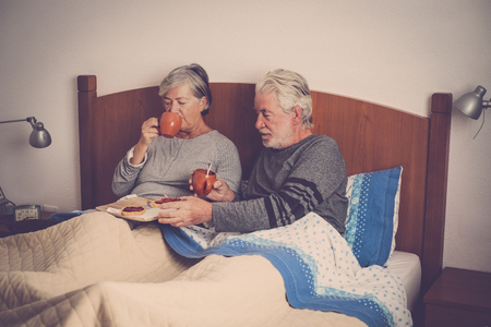 Old senior mature caucasian couple enjoy breakfast in the morning at bed in the bedroom at home - retired people live together and eat healthy food to start the day with energyの写真素材