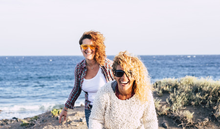 Happy cheerful laughing beautiful couple of people women outdoor having fun in friendship together enjoying the outdoor leisure activity with blue ocean and sky in backgroundの写真素材
