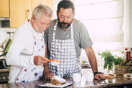 Family at home at work in the kitchen with senior mature father and adult son preparing a cake together like friends - diversity and mixed generations in the houseの写真素材