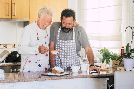 Caucasian couple at home preparing a cake together - father and son adult and senior working in the kitchen - cooking men chef at work with joy and happinessの写真素材