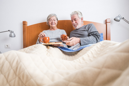 Old senior mature caucasian couple enjoy breakfast in the morning at bed in the bedroom at home - retired people live together and eat healthy food to start the day with energyの写真素材