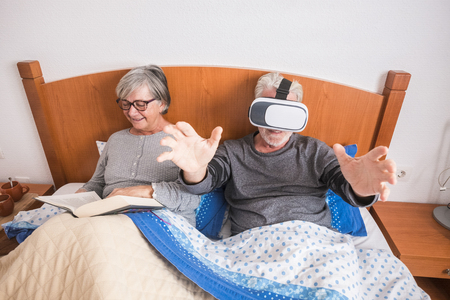 Happy alternative funny old senior couple of caucasian people grandfathers enjoy in the berdroom - man with goggles headset play virtual games and woman read a bookの写真素材
