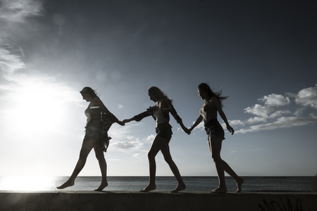 Team and cooperation concept with friends people group beautiful girls walking together balanced on a wall with blue ocean and sunset in background - outdoor leisure activityの写真素材
