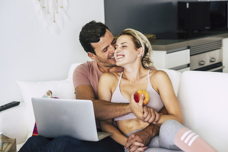 Cheerful and happy couple hugging and laughing at home lay down on the sofa - love and relationship concept with lifestyle healthy concept - technology and home working for young peopleの写真素材