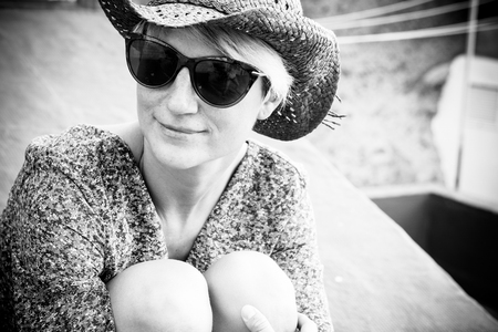 Beautiful black and white romantic portrait with blonde young caucasian woman sitting and hugging her nkees outdoor with sunglasses and little smileの写真素材