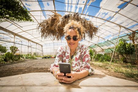 Beautiful cheerful curly blonde young caucasian woman use modern phone typing message or searching on internet in outdoor rural scenic place during summer - people and technology conceptの写真素材