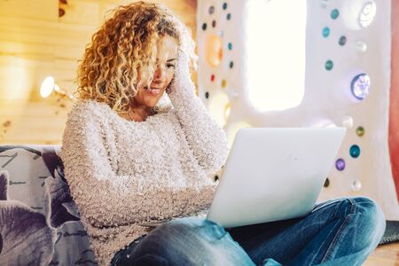 Beautiful curly blonde adult woman use computer laptop in the bedroom at home or hotel - enjoying free office job and freelance nomad lifestyle - people and technology for lifestyleの写真素材