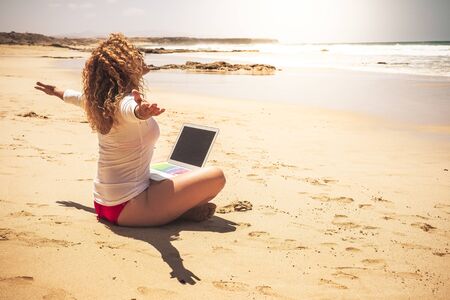 Happy free people caucasian woman sit at the ebach with computer laptop on her legs opening arms enjoying the freedom for vacation summer and for work free from stress office in the cityの写真素材