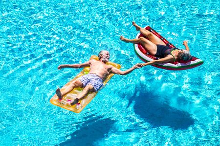 Happy relaxed people caucasian senior adult couple taking hands lay down with trendy coloured lilos on a blue water swimming pool during a summer holiday vacation - enjoying active retired lifestyleの写真素材