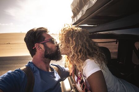 Adult caucasian cute couple of traveler kiss each other with sun in backlight - car traveling together with love and relationship - sandy desert in backgrounf and bright sky - man with beardの写真素材
