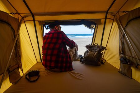 Lonely hipster traveler sitting inside a tent in free camping at the beach - relax and travel concept with alternative backpacke rlifestyle - wanderlust photographer looking the landscapeの写真素材