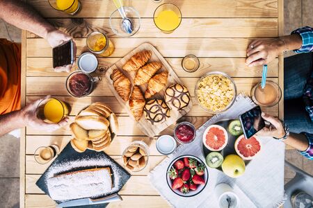 Above top view of wooden table with couple having nice breakfast in the morning with tasty cakes and fruit for different lifestyle concept - people with technology addiciton conceptの写真素材