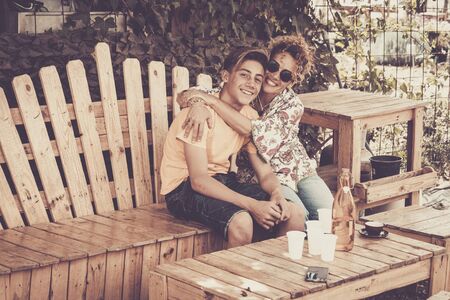 mother and son hugging with smile and enjoy leisure time together with love. family teenager boy 14 years old and mom 43 have fun sitting on a wodd bench outdoor in a sunny day. happiness family conceptの写真素材