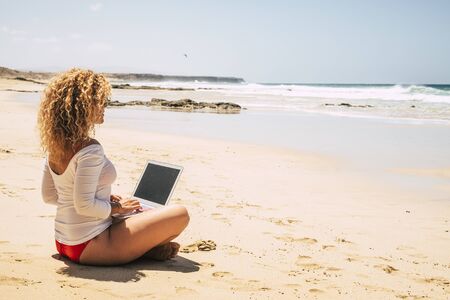 Concept of people work free from office - modern digital nomad lifestyle working in technology related jobs - happy women in bikini using laptop at the ebach enjoying the ocean viewの写真素材
