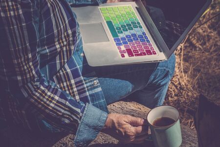 Travel and technology concept for modern mature senior people - hipster retired woman sit down with coloured computer laptop in the nature with a cup of coffeeの写真素材