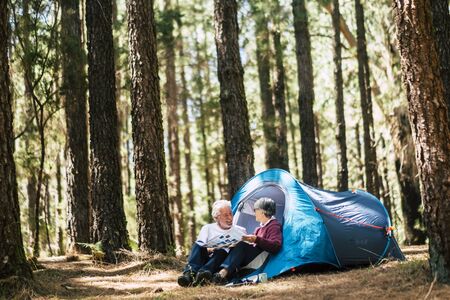 Active people old aged caucasian senior couple travel with camping tent - sit down in the forest having fun together - relationship forever concept for traveler wanderlust retired man and womanの写真素材