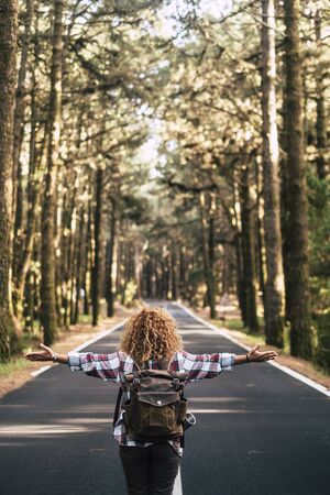 Alternative travel concept with curly hipster woman viewed from back open his arms and feel the freedom of the outdoors nature standing in the middle of a ling road - happiness and wanderlust lifestyleの写真素材